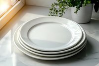 Stack of clean white ceramic dinner plates on marble countertop with natural sunlight and green plant decoration, minimalist kitchen setting.