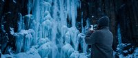 Photographer in winter gear capturing frozen waterfall landscape with icicles and snow-covered rocks in cold climate environment.