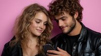 Young couple looking at smartphone together, sharing intimate moment against pink background. Caucasian woman with curly hair and man in leather jacket smiling.