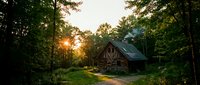 Rustic wooden cabin nestled in dense forest with golden sunlight filtering through tall trees at sunset, surrounded by lush green vegetation and natural woodland landscape.