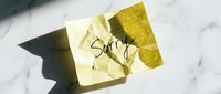 Crumpled yellow sticky note with handwritten word Sorry on white marble surface in sunlight, conveying apology and regret.