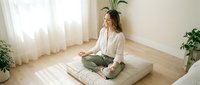 Woman in casual clothing sitting cross-legged on floor cushion in minimalist room with plants and natural light, practicing meditation or mindfulness for wellness and relaxation.