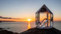 Crystal prism house sculpture on rocky coastline at golden sunset with ocean waves and warm sky colors creating peaceful meditation scene.