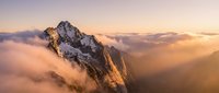 Majestic snow-capped mountain peak emerging through golden clouds at sunrise, dramatic alpine landscape with warm light illuminating rocky cliffs and misty valleys.