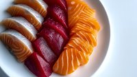 Fresh raw salmon and tuna sashimi slices arranged in rows on white ceramic plate, showing vibrant orange and red colors with distinctive fish texture patterns.