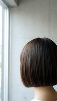 Woman with short dark brown bob hairstyle standing by bright window in minimalist interior space, rear view showing sleek hair texture and modern cut for salon, beauty, and hair care concepts.