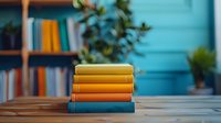Stack of colorful hardcover books on wooden table against turquoise wall with blurred bookshelf and plant in background, creating cozy reading atmosphere.