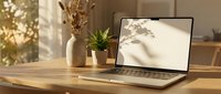 Laptop with blank white screen on wooden desk, warm sunlight, plants and vase in background, cozy home office workspace mockup.