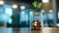 Young plant growing from coins in glass jar against blurred office background, representing financial growth, investment and wealth accumulation concept.