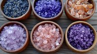 Colorful bath salts in wooden bowls arranged in rows, featuring pink, purple, and blue crystals for aromatherapy and spa treatments on dark wooden surface.