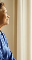 Senior woman in blue shirt gazing thoughtfully toward window with soft natural light, conveying contemplation and peaceful reflection for wellness and lifestyle content.