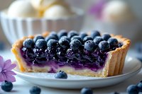 Fresh blueberry tart with purple filling and golden crust on white plate, garnished with fresh berries and purple flowers, close-up food photography.
