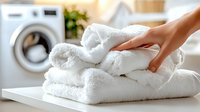 Fresh white fluffy towels stacked on table with washing machine in background, female hand touching soft clean laundry in bright modern bathroom.