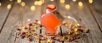Small glass bottle with orange liquid and cork stopper surrounded by colorful dried flowers on rustic wooden surface with warm bokeh lights background.