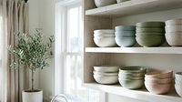 Organized kitchen shelves displaying stacked ceramic bowls in neutral and sage tones, with fresh greenery in vase by window, suitable for home decor and interior design projects.