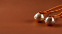 Traditional metal bells with ornate pattern on brown background, attached to orange cord, used in meditation practices and spiritual ceremonies.