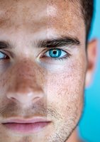 Close-up portrait of young Caucasian male with intense blue eyes, freckles, and stubble against turquoise background, showing half of face in dramatic lighting.