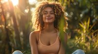 Young woman with curly hair smiling in golden sunlight, surrounded by lush tropical foliage, creating warm and peaceful atmosphere for wellness and lifestyle content.