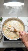 Creamy chicken soup with vegetables cooking in pot on stove with steam rising under modern kitchen hood while hand stirs with wooden spoon.
