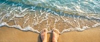Feet standing on sandy beach with crystal clear turquoise ocean waves washing ashore creating white foam patterns on golden sand for summer vacation relaxation.