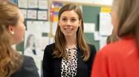 Professional female teacher with long brown hair smiling while speaking with colleagues in modern classroom setting with educational materials and colorful displays.