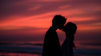 Romantic couple silhouette kissing at sunset beach with dramatic pink and orange sky clouds creating intimate moment for wedding or love concept.
