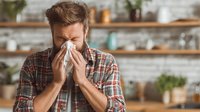 Man with cold blowing nose into tissue in cozy kitchen setting with wooden shelves and plants in background.
