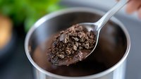 Crushed dark chocolate pieces on metal spoon over black coffee cup, macro shot showing rich texture and melting chocolate bits for food photography.