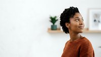 Young African American woman with curly black hair wearing rust-colored sweater, looking upward thoughtfully in bright modern interior space with shelving and plants.