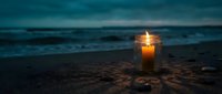 Glowing candle in glass jar on sandy beach at twilight with ocean waves in background creating peaceful atmospheric mood for meditation and relaxation.
