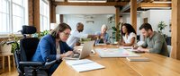 Diverse business team collaborating in modern office meeting room with laptops and documents on wooden table during productive work session.