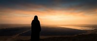 Silhouetted figure standing on mountain peak at sunset, gazing at golden sky and distant landscape below, conveying solitude, contemplation and adventure for travel and lifestyle content.