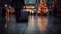 Traveler with luggage walking through airport terminal decorated with Christmas trees and festive lights, creating warm holiday atmosphere.