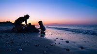Father and young child building sandcastle together on beach during golden hour sunset with colorful sky and ocean waves creating family bonding moment.