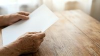 Senior adult with brown skin tone holding blank white card on wooden table surface, suitable for greeting cards, invitations, announcements, and business messaging.