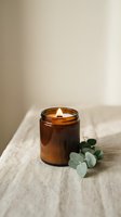 Lit scented candle in amber glass jar with eucalyptus leaves on linen fabric, creating a calming spa and wellness atmosphere for relaxation and home decor.