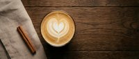 Cappuccino with heart latte art on rustic wooden table next to cinnamon stick and linen napkin for cafe menu design and coffee shop marketing materials.