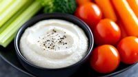 Creamy white dip garnished with black pepper served with fresh vegetables including celery stalks, cherry tomatoes, and carrots on dark plate for healthy snacking.