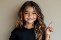 Happy young girl with long wavy brown hair pointing finger up, smiling cheerfully in black t-shirt against neutral beige background for advertising campaigns.
