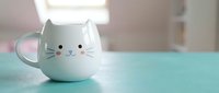 Adorable white cat-shaped ceramic mug with pink ears and whiskers sits on turquoise surface in bright kitchen setting for morning coffee or tea enjoyment.