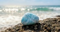 Blue larimar gemstone resting on rocky shore with turquoise ocean waves and sandy beach in soft focus background for wellness and meditation concepts.