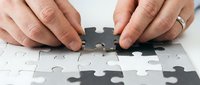 Hands placing final puzzle piece to complete white jigsaw puzzle on table, symbolizing problem solving, teamwork and business solutions.