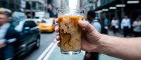 Caucasian male hand holding iced cola beverage on urban street with yellow taxi and pedestrians blurred in background, suitable for beverage marketing and lifestyle content.