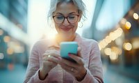 Mature woman with glasses and gray hair smiling while using smartphone in urban setting with bokeh lights at sunset.