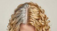 Woman showing gray roots growing out from blonde curly hair demonstrating natural aging process and hair color maintenance needs for beauty salon services.