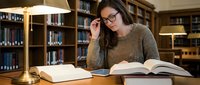 Young woman with glasses studying in library, reading books at wooden desk with warm lighting, academic research and education concept.