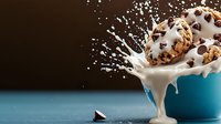 Chocolate chip cookies splashing into milk with dramatic droplets and motion capture against dark background in blue ceramic bowl. Food photography.