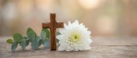 Wooden cross with white chrysanthemum flower and eucalyptus leaves on rustic surface, soft bokeh background, funeral or Easter memorial concept.