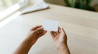 Woman with medium skin tone holding blank white business card or paper mockup on wooden desk with plant in background, suitable for branding and corporate presentations.
