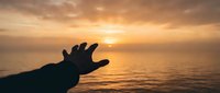 Silhouette of hand reaching toward golden sunset over calm ocean waters creating peaceful moment of connection with nature and tranquil evening atmosphere.